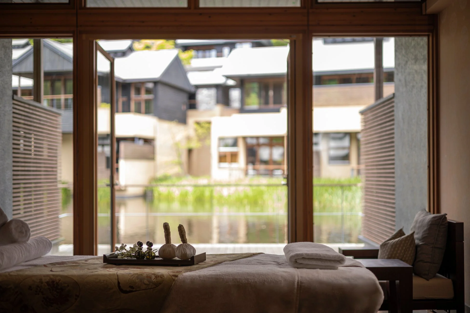 マッサージベッドが置かれたスパルームの様子。窓からは水辺と趣のある建物が見え、部屋にはハーブボールなどが準備されており、癒やしとリラックスを感じさせる空間です。
