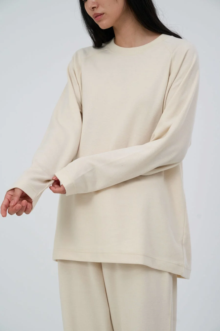 白い背景の前で、ベージュの長袖トップスとボトムスを着用した女性が、片方の袖を軽く掴んでいる様子を捉えた画像です。シンプルでリラックスした雰囲気のファッションアイテムが強調されています。