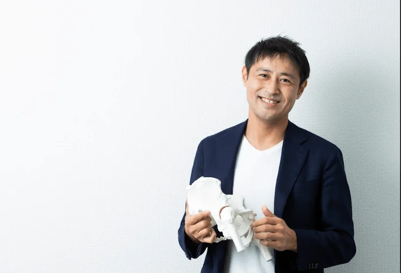 白背景の前で笑顔の男性が骨盤の模型を持っている。医療や健康分野の専門家や教育者を連想させるイメージ。