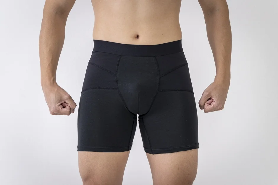 白い背景の前で、黒いボクサーパンツまたはコンプレッションショーツを着用した男性が、両手を握りしめて立っている様子を捉えた画像です。