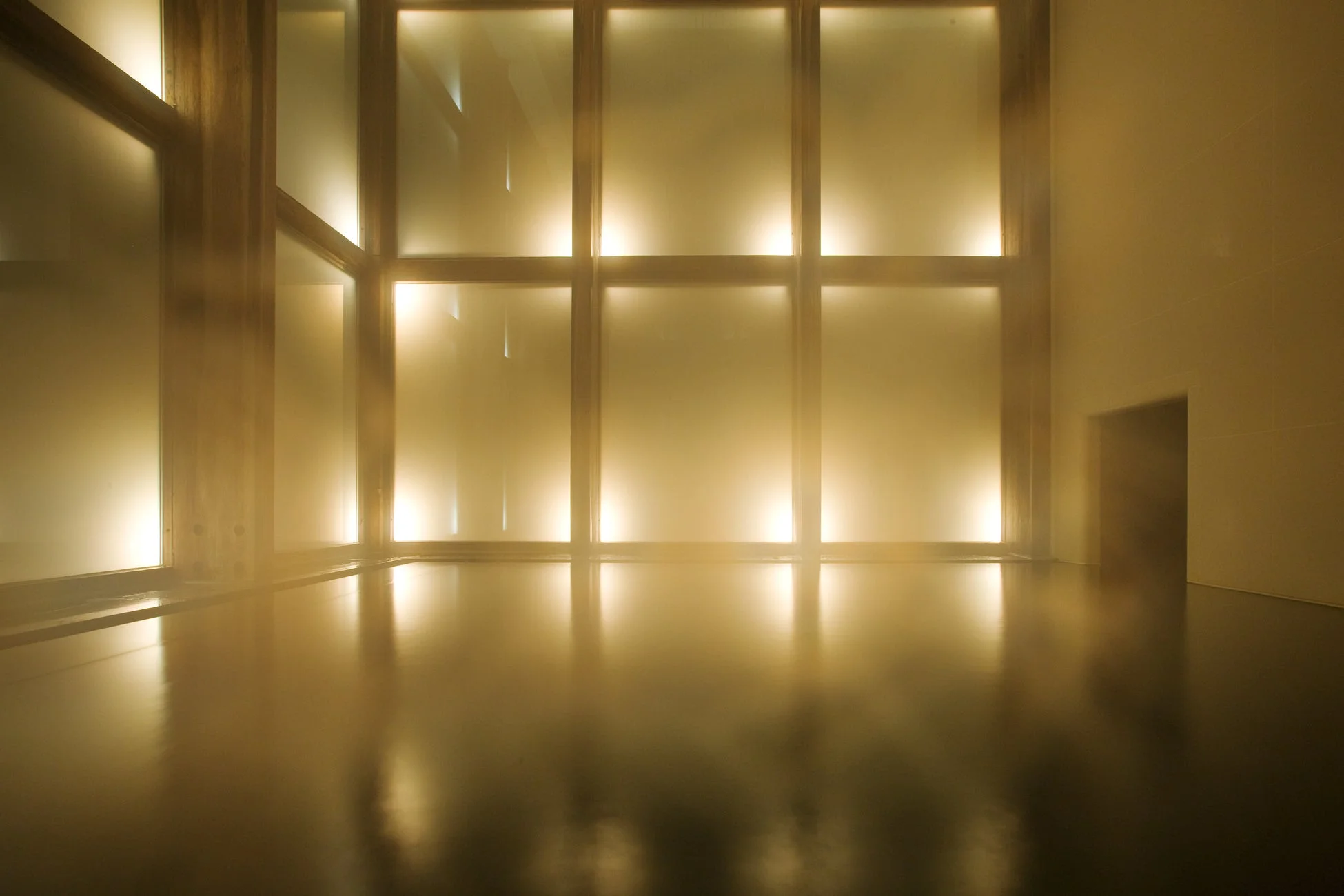 暖かく黄金色の光に包まれた広々とした室内空間。木枠の窓から差し込む柔らかな光が反射する床が印象的で、静かで幻想的な雰囲気を醸し出している。右端には暗い扉が見える。