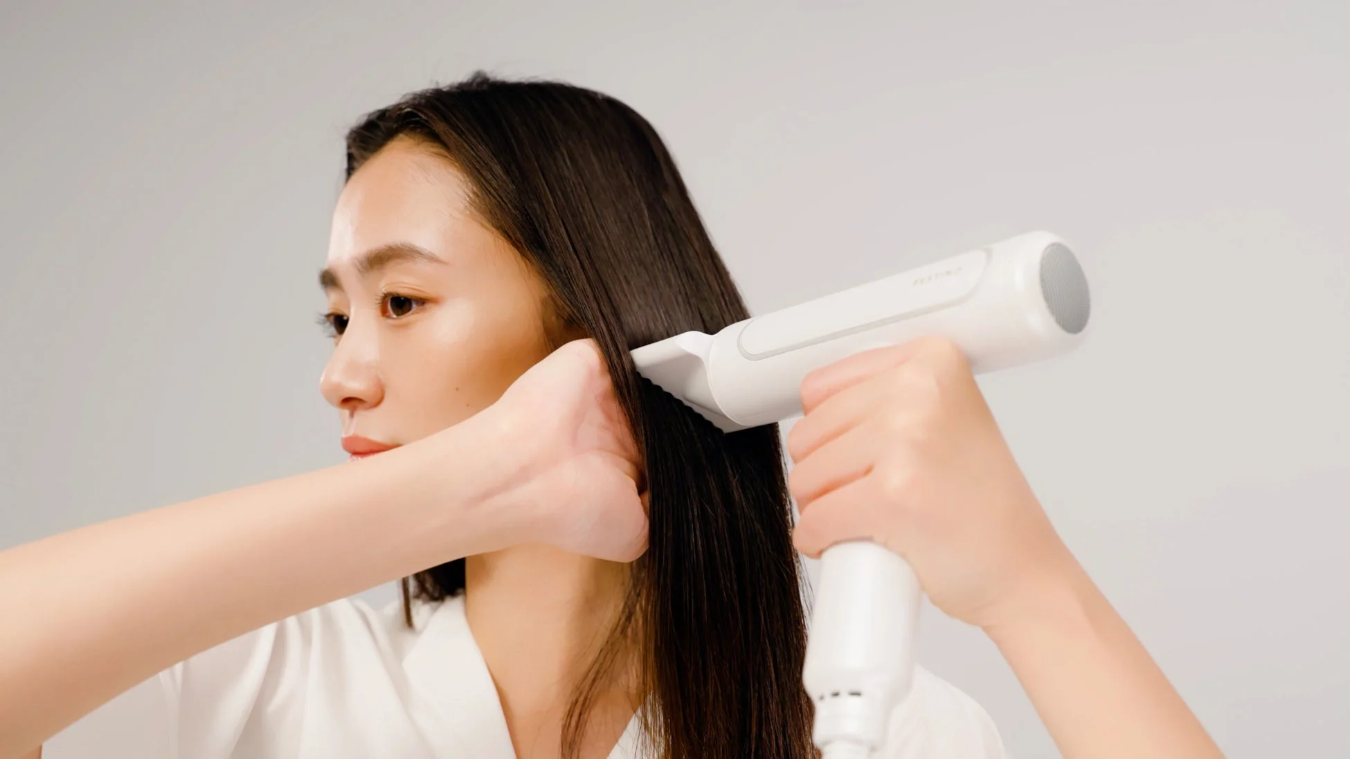 白い背景の前で、女性が白いヘアスタイリングデバイスを使って髪を整えている様子