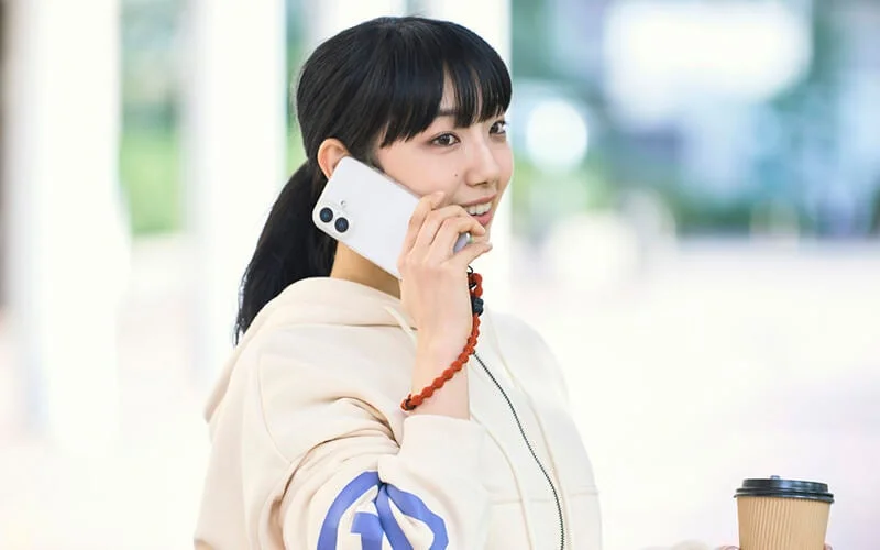 ポニーテールの若い女性が笑顔でスマートフォンで通話しており、もう片方の手にはコーヒーカップを持っている