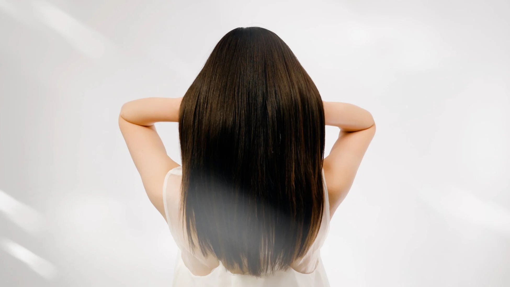 白い背景を背に、長い黒髪の女性が後ろ姿で写っています。両腕を頭の後ろに回し、美しいストレートヘアが強調されています。