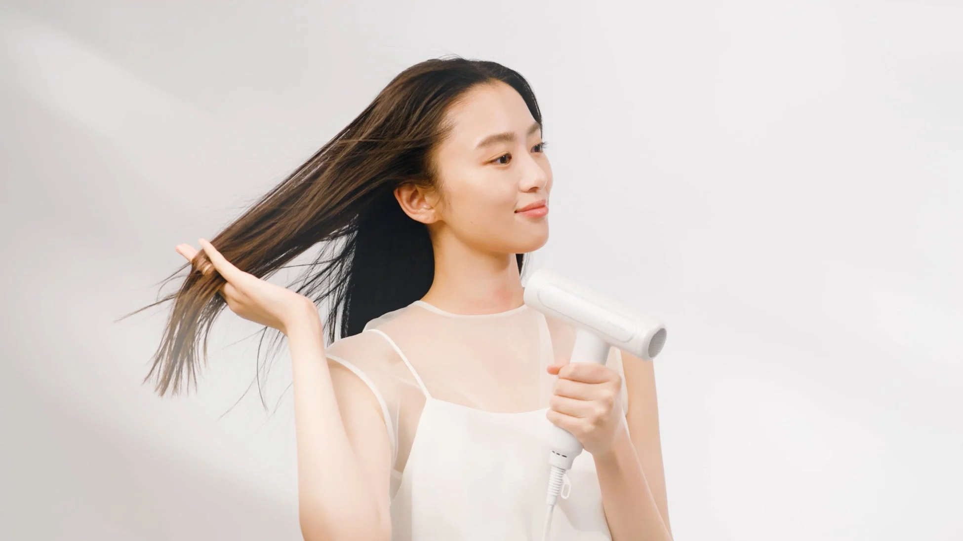 白い背景の前で、一人の女性が白いドライヤーを使って長い髪を乾かしている様子