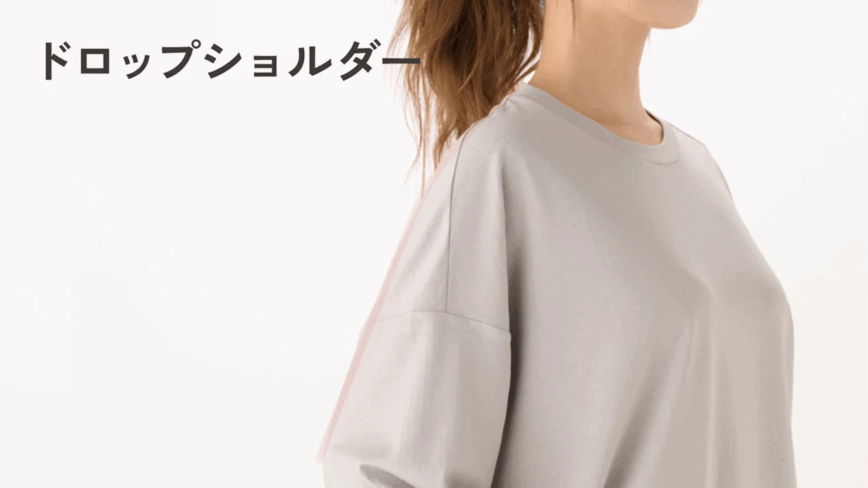 白い背景の前で、ドロップショルダーのライトグレーのトップスを着た女性の上半身が写っています。