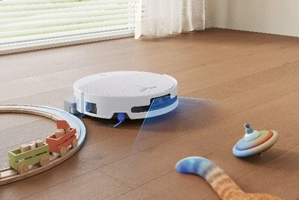 木製の床の部屋で、白いロボット掃除機が稼働しており、周囲には木製のおもちゃの列車や猫のしっぽが見える。明るいリビングの日常風景。