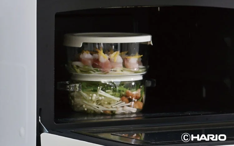 電子レンジの中に、HARIO製のスタッキングできる蒸し料理用容器が置かれ、肉巻きと野菜が調理される準備が整っている様子。ヘルシーな食事が手軽に作れることを示唆しています。