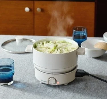 白い電気鍋が食卓に置かれ、湯気を立てながら野菜が調理されている様子を捉えた画像です。