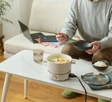 自宅のリビングで、一人がローテーブルで電気鍋を使いながら食事をしている様子です。