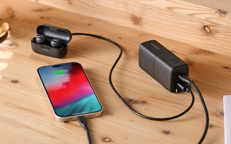 木目のデスク上で、スマートフォンとワイヤレスイヤホンが黒い充電器に接続され、同時に充電されている様子