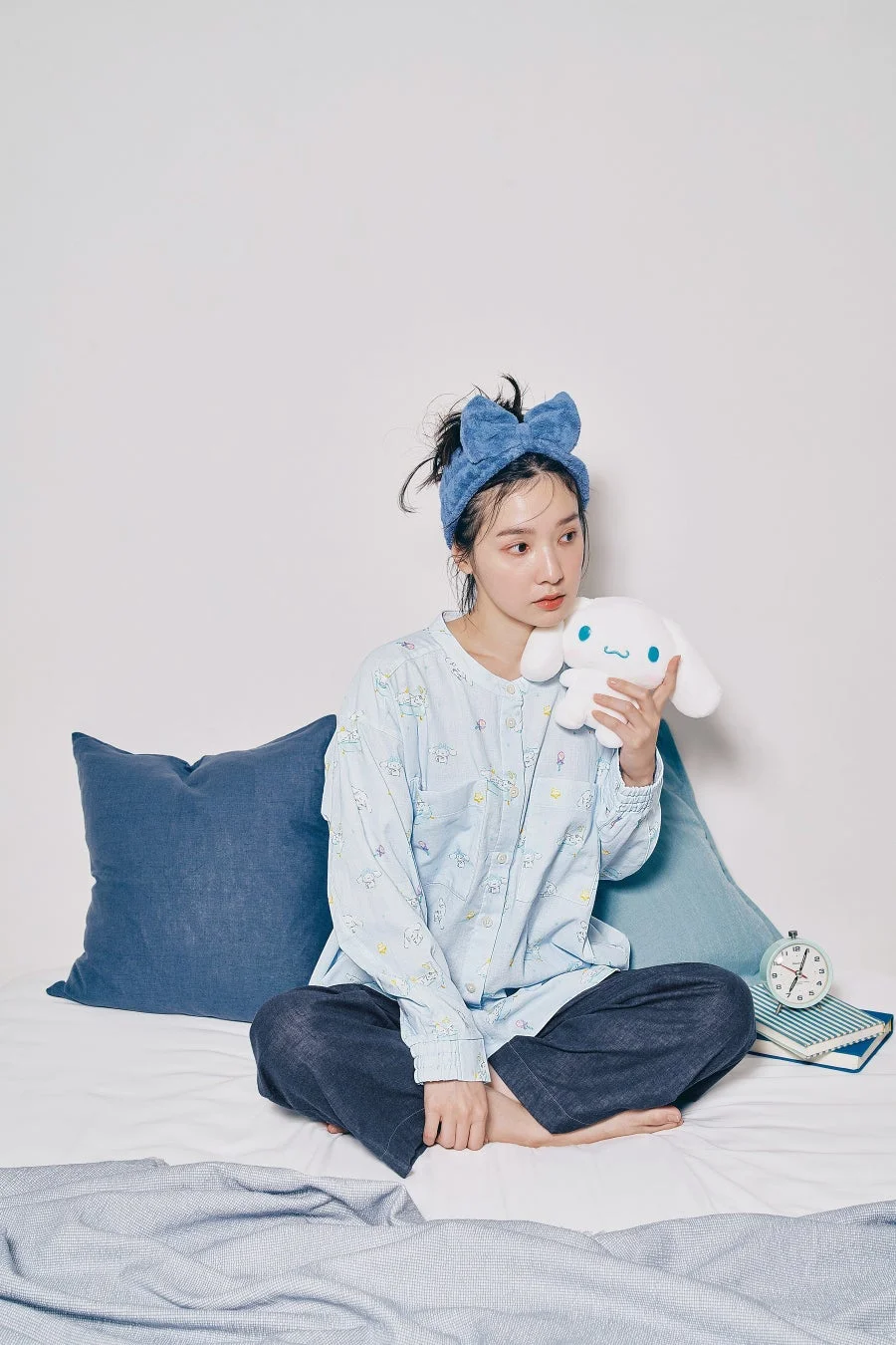 ベッドの上で青いパジャマとヘアバンドを身につけた女性が、シナモロールのぬいぐるみを抱えて座っている