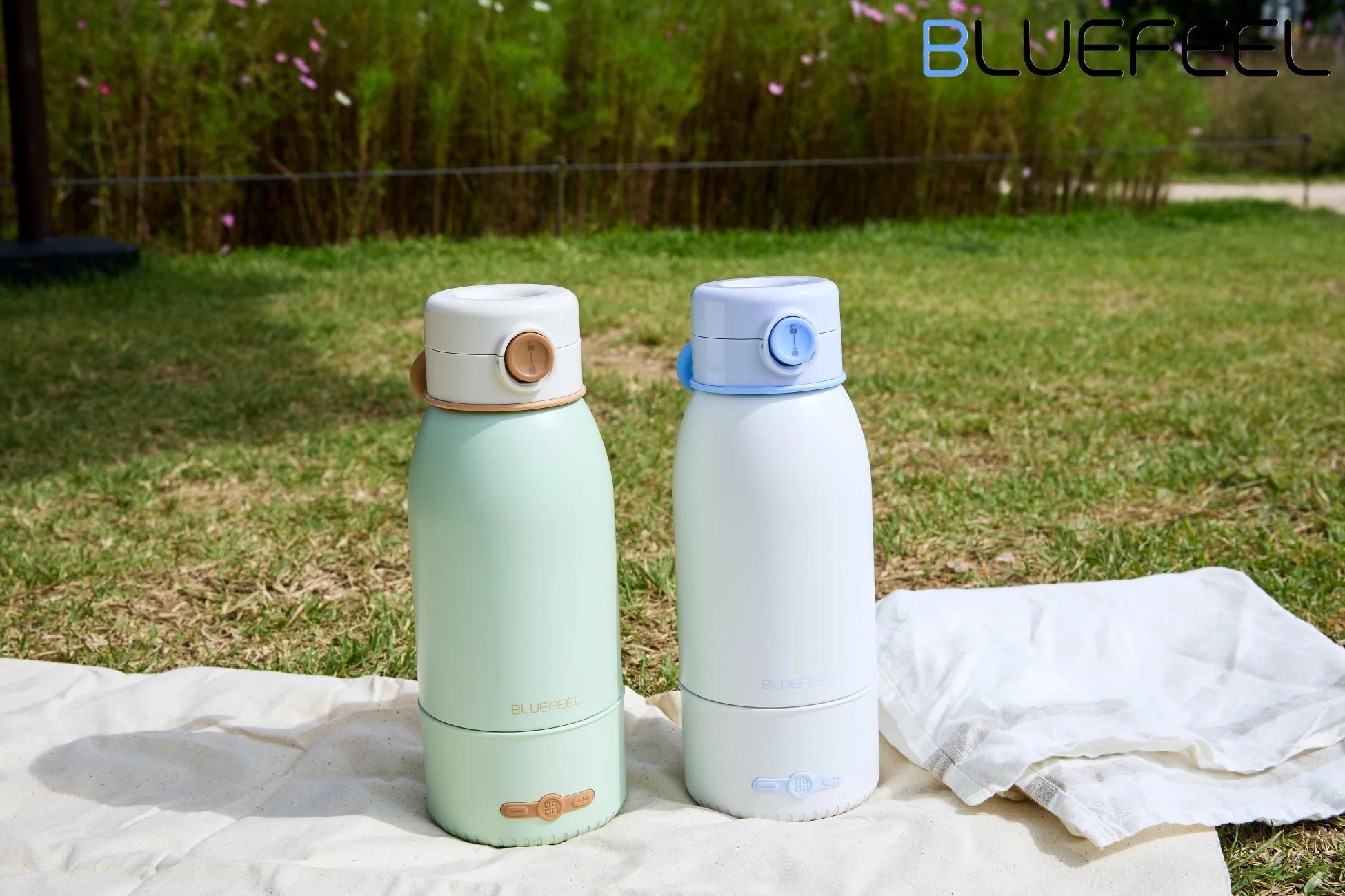 芝生の上に敷かれた白い布の上に、ミントグリーンと白の2つのポータブルブレンダーが置かれています。背景には緑豊かな植物が広がり、のどかなアウトドアの雰囲気を醸し出しています。