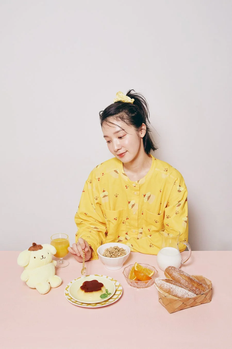黄色のパジャマを着た女性が、ポムポムプリンのぬいぐるみと一緒に、プリンやシリアルなどを用意したピンクのテーブルで朝食を楽しんでいる
