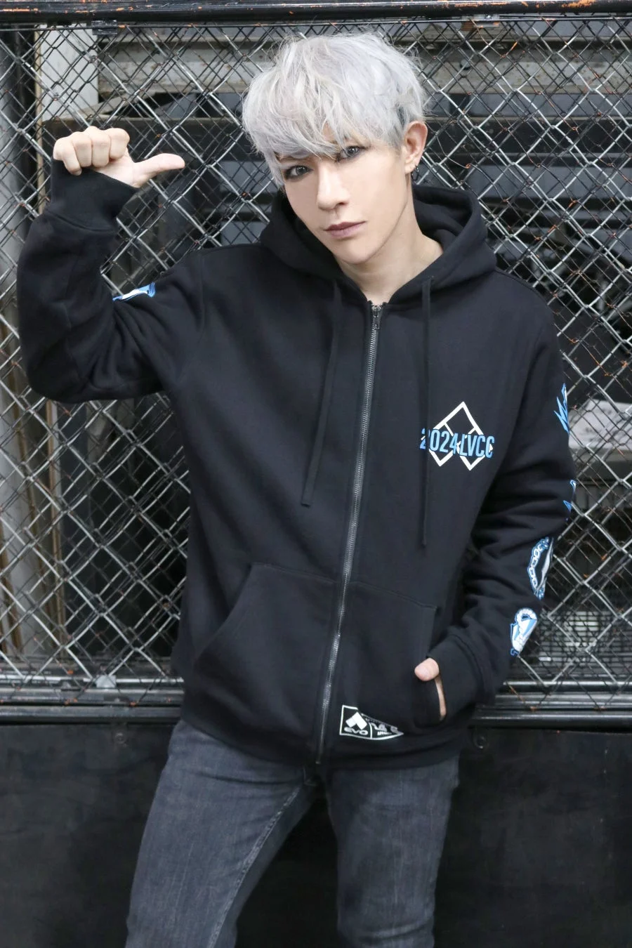 銀髪の男性が黒いジップアップパーカーとジーンズを着用し、ワイヤーフェンスの前でポーズをとっています。パーカーには青いロゴがデザインされており、クールな印象です。