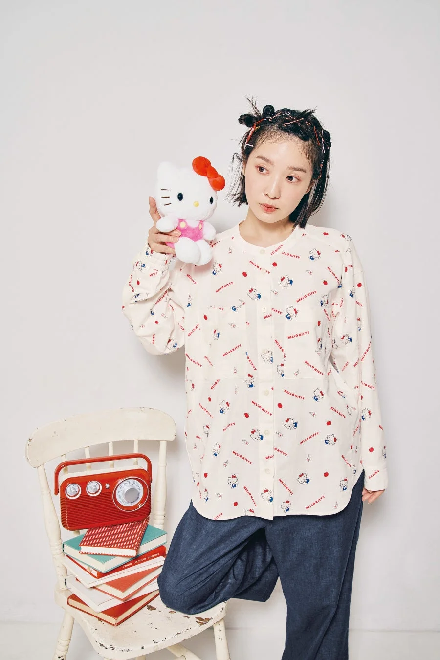 ハローキティ柄のシャツを着た女性が、ハローキティのぬいぐるみを持ち、ヴィンテージラジオと本が積まれた椅子と共にポーズ