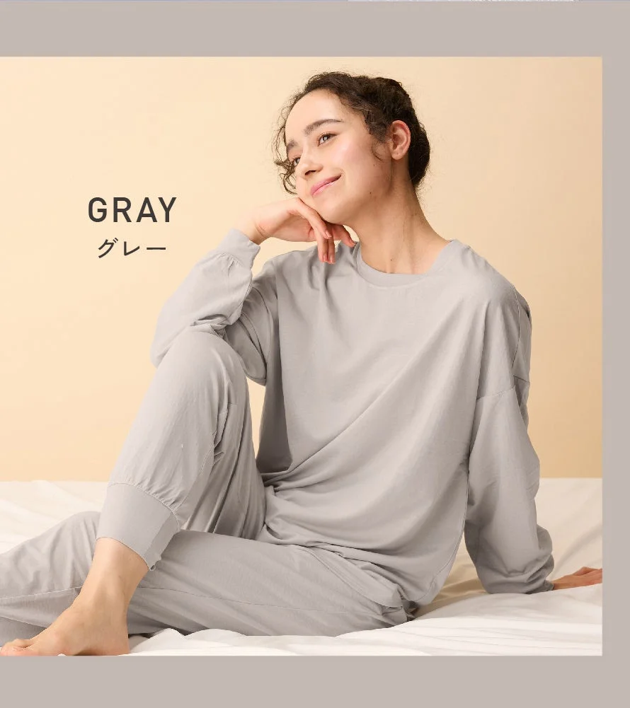 グレーのルームウェアを着た女性がベッドに座り、リラックスした笑顔を見せています。