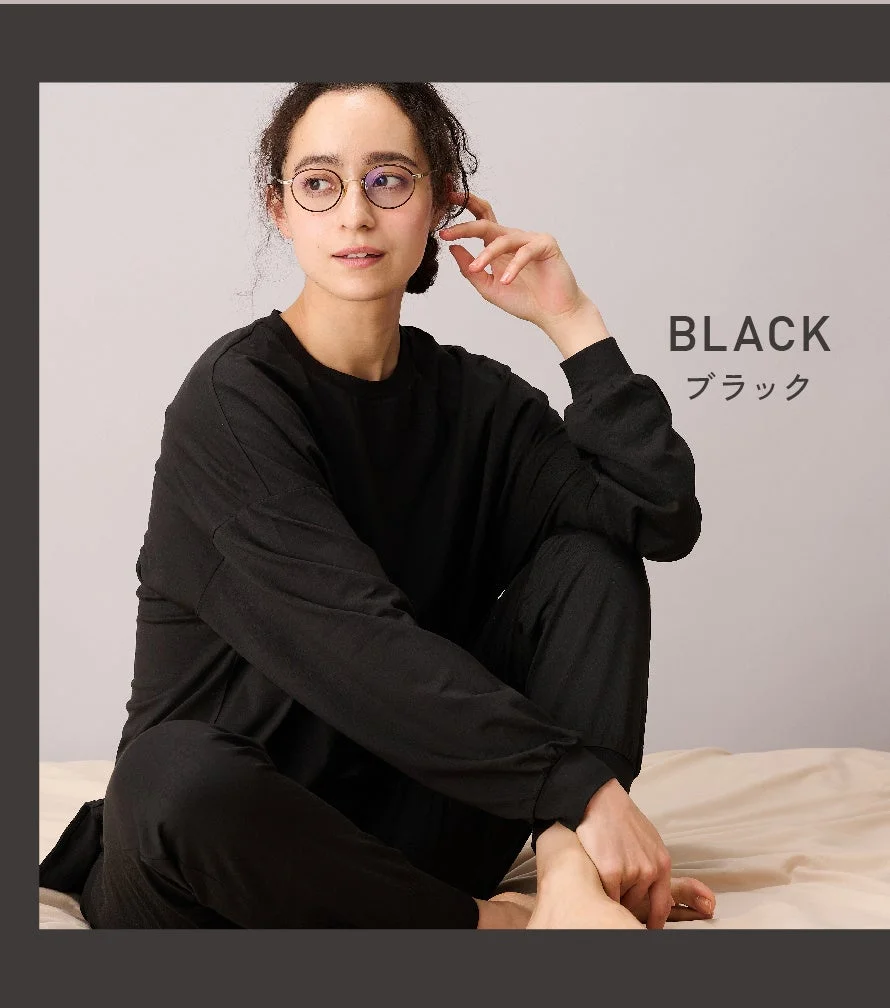 ベッドに座り、黒いルームウェアとメガネを着用した女性がリラックスした様子で写っています。