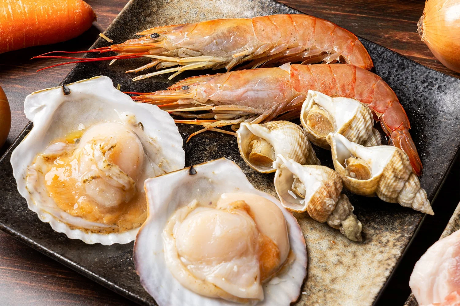 新鮮なエビ、ホタテ、サザエなどの魚介類が黒い皿に盛り付けられています。奥にはニンジンや玉ねぎも見え、豪華な海鮮料理の食材であることが伺えます。