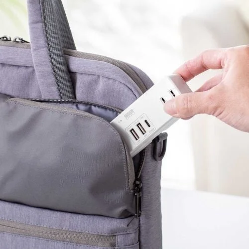 グレーのビジネスバッグのポケットに、SANWA SUPPLYのロゴが入った白いUSBポート付き電源タップを収納している様子です。持ち運びに便利なコンパクトな電源タップで、旅行や出張に役立つアイテムです。