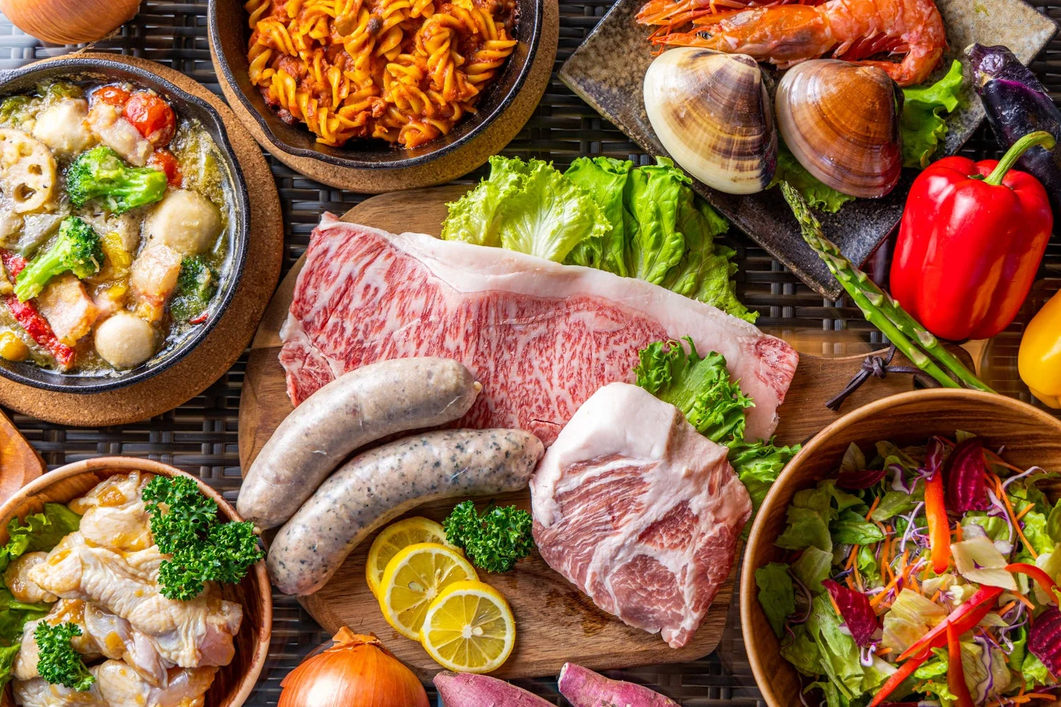 豪華な肉、魚介類、新鮮な野菜が豊富に並べられた食卓の俯瞰写真。和牛やソーセージ、エビ、アサリ、パスタ、鍋料理、サラダなど、多彩な料理の準備風景を捉えている。