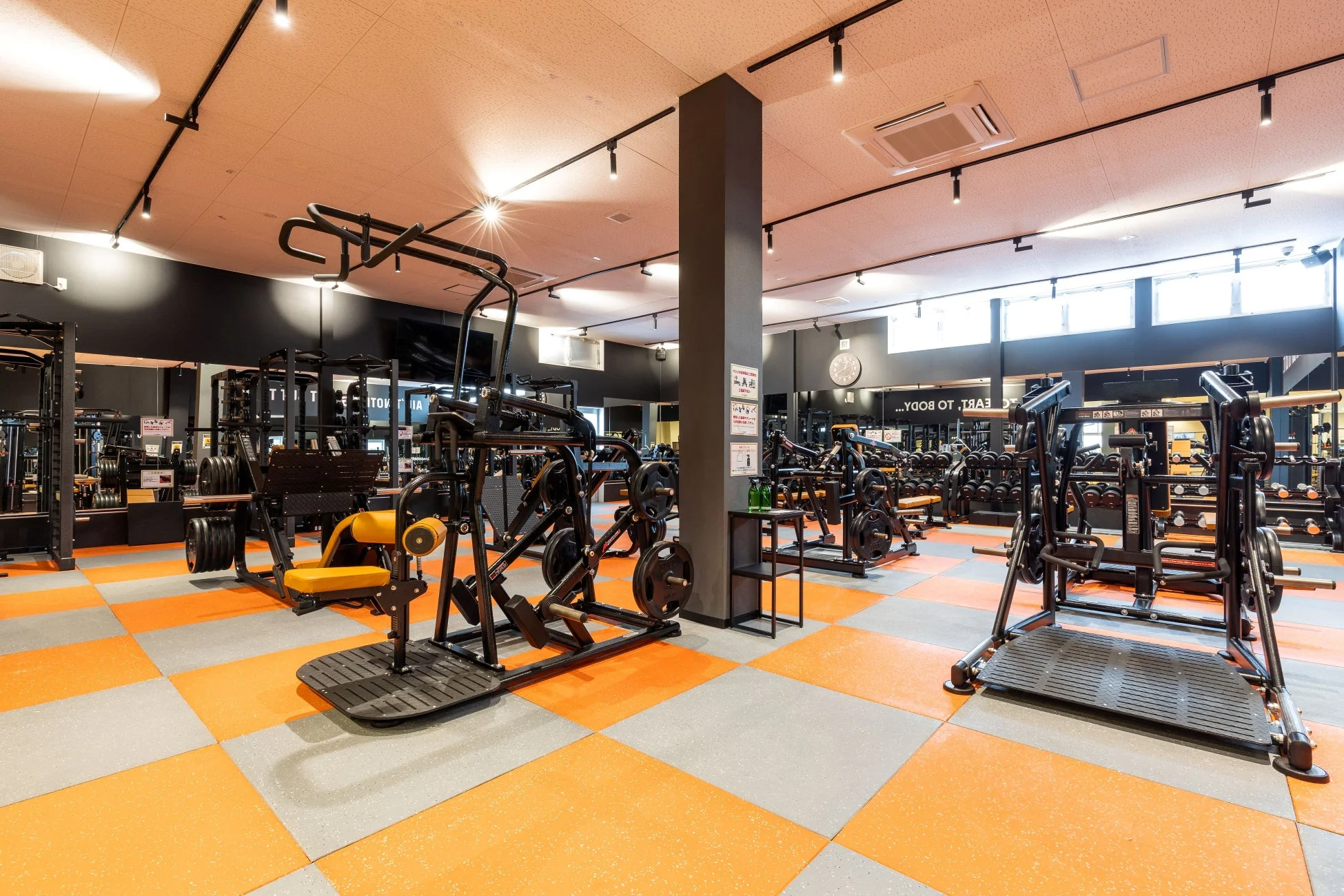 オレンジとグレーの市松模様の床が特徴的な、広々としたトレーニングジムの内部です。様々な種類の筋力トレーニングマシンが整然と並べられており、清潔感のある現代的なフィットネス空間が広がっています。