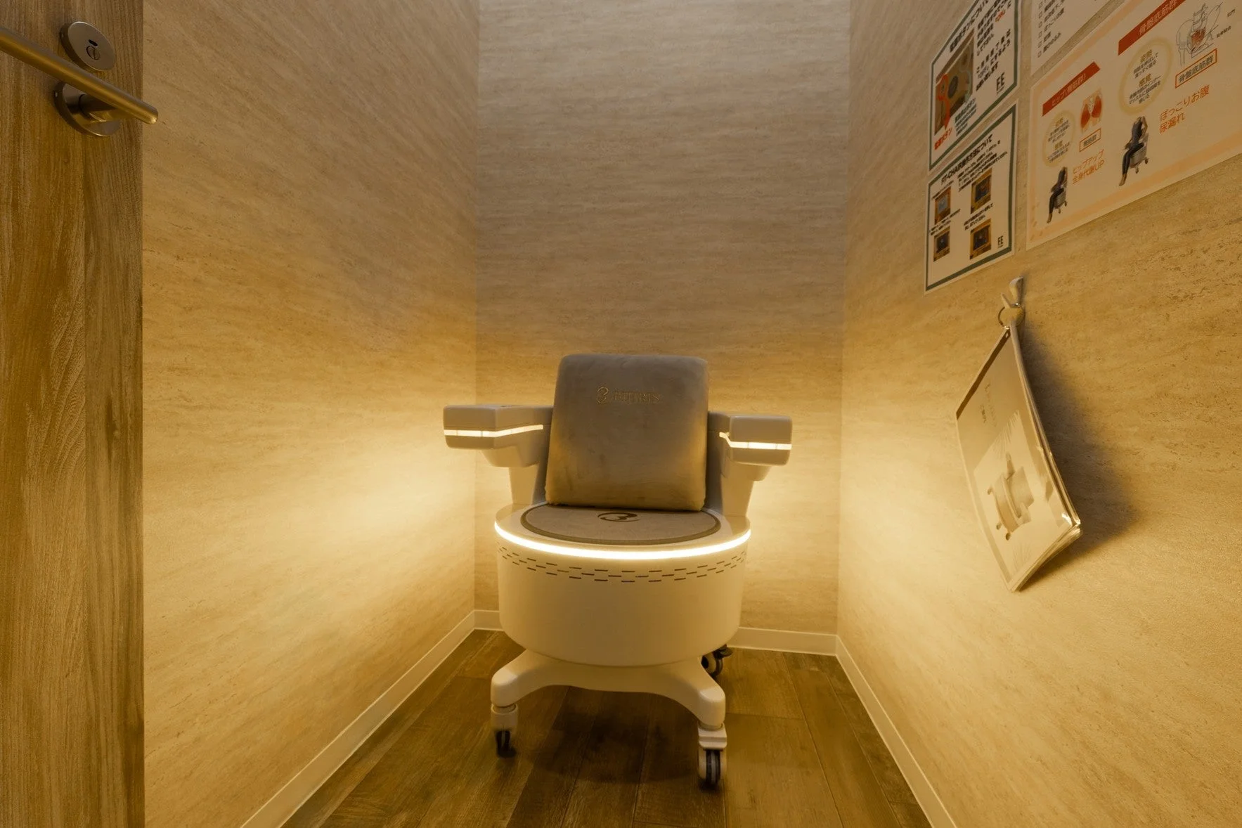 暖色系の照明が特徴的な個室に設置された、未来的なデザインの白色高機能チェア。壁のポスターから骨盤底筋トレーニングや美容、全身ケアに関連する医療・健康機器であることが伺える。