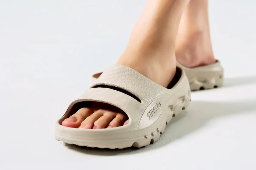 白い背景で、人物がベージュ色のスライドサンダルを履いている足元をクローズアップした画像です。快適そうなデザインが特徴です。