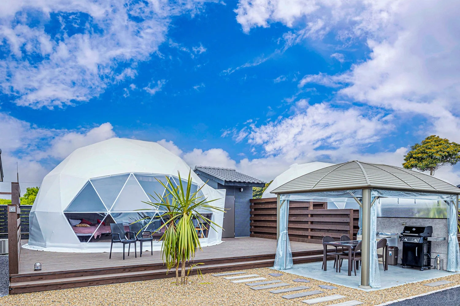 青い空と白い雲の下、ドーム型テントが並ぶグランピング施設です。ウッドデッキや屋外のバーベキュー設備を備えたガゼボがあり、リラックスできる宿泊体験を提供しています。