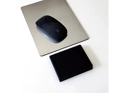 黒いApple Magic Mouseがシルバーのマウスパッドの上に置かれ、その手前には黒い四角い物体がある