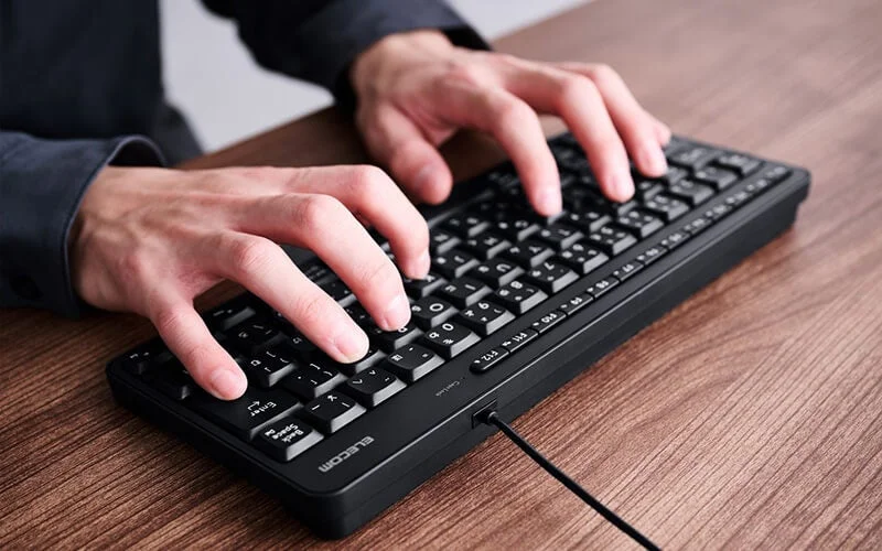 木目調のデスクで、黒いキーボードを両手でタイピングしている人物