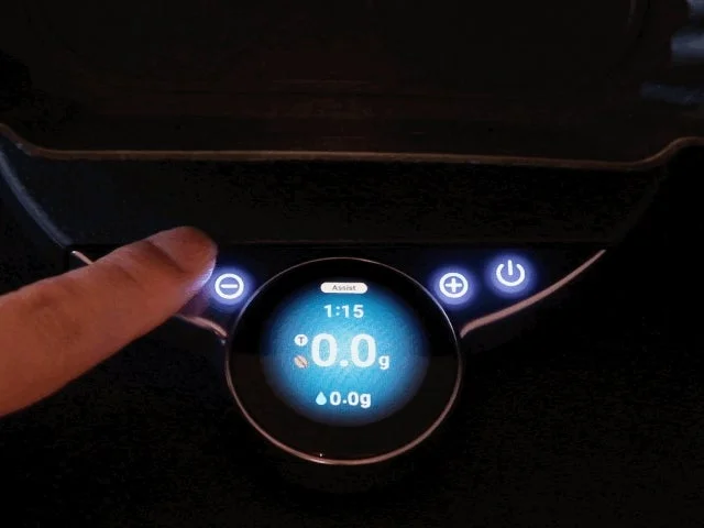 デジタル計量器のボタンを押している様子。ディスプレイには「Assist」や「1:15」、「0.0g」と表示されており、コーヒー豆や水の量を測るスマートスケールであると推測される。