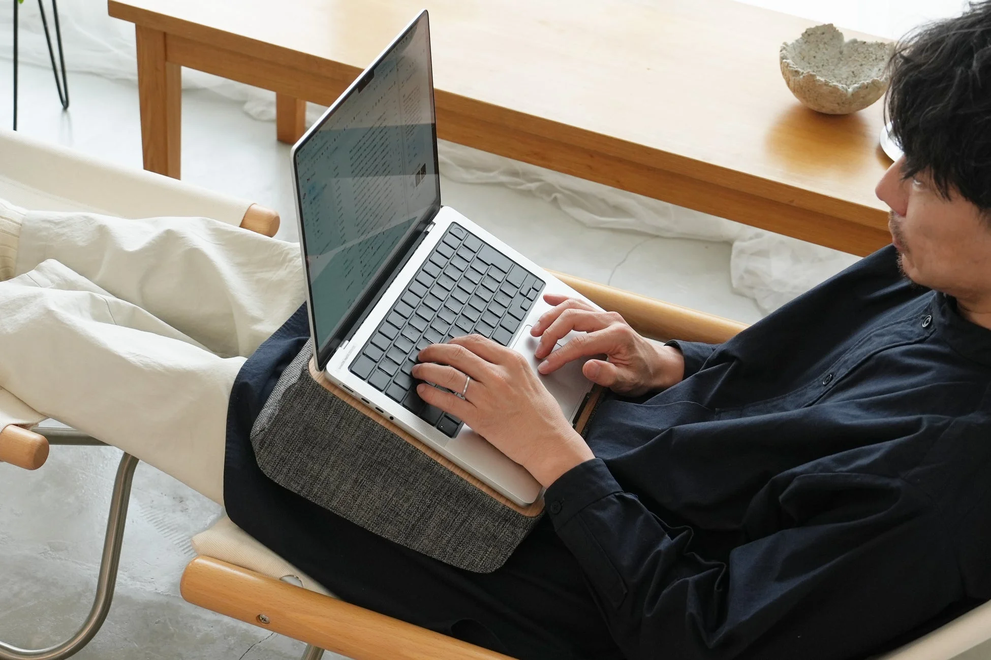 男性がリラックスした姿勢で椅子に座り、ラップデスクに置いたノートパソコンで作業している様子。自宅での快適なワークスタイルを思わせる一枚で、背景には木製のテーブルが見えます。