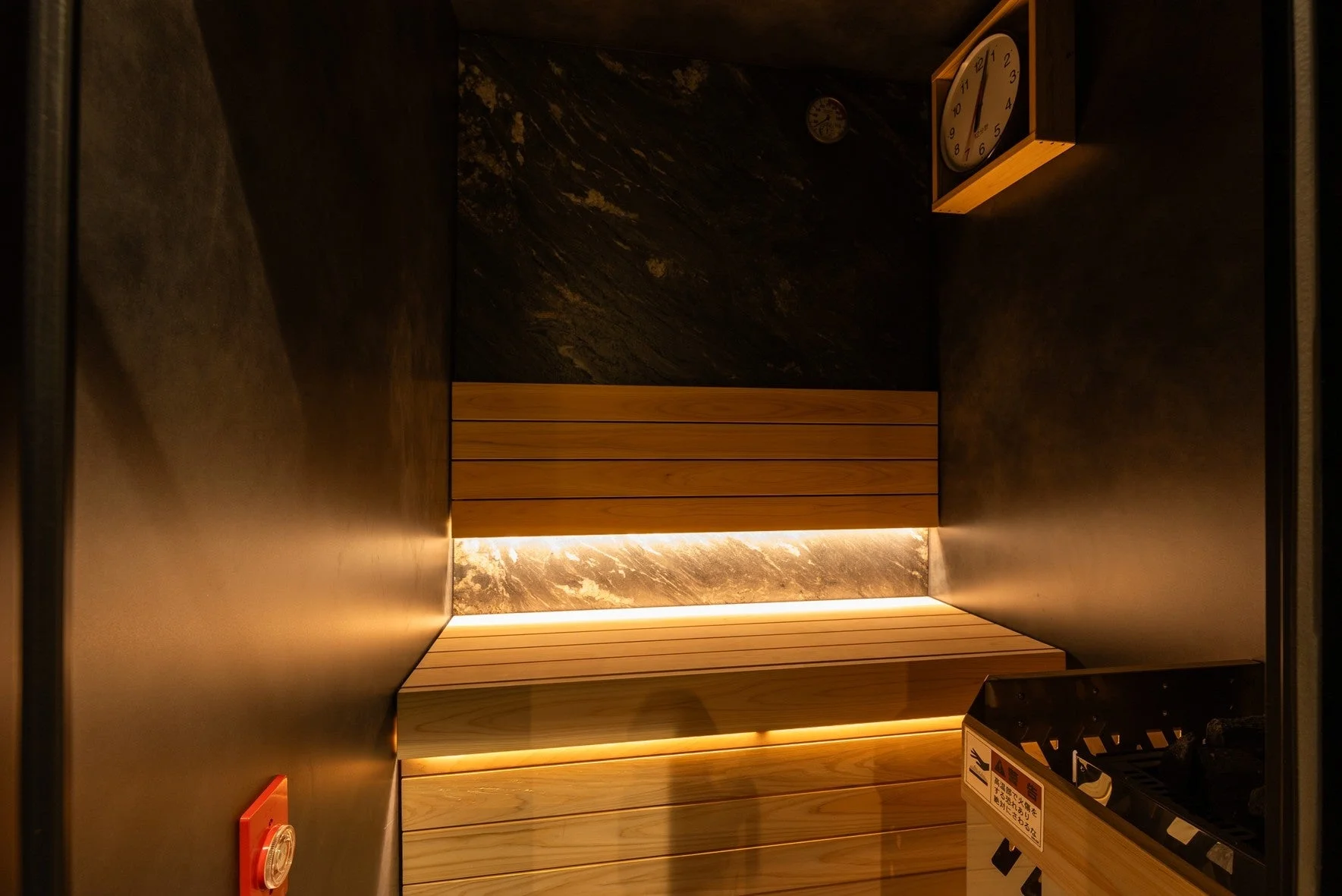 間接照明が温かく照らすモダンなサウナ室の内部。木製ベンチと黒い壁が特徴的で、リラックスできる空間を演出しています。壁には時計、右下にはサウナヒーターがあります。