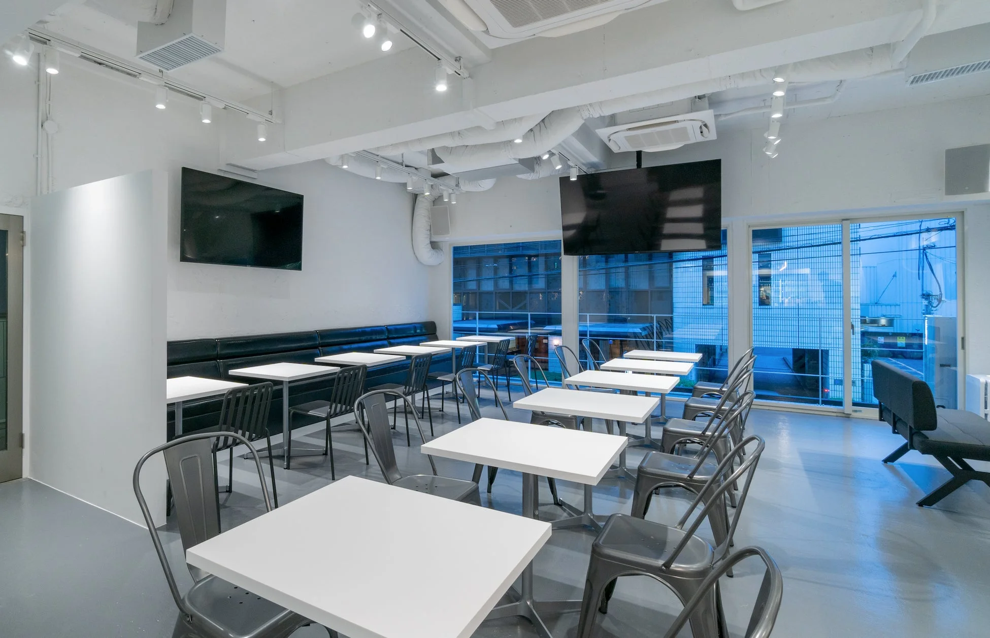 白を基調としたモダンでシンプルなカフェや飲食店の内装です。複数のテーブルと椅子が並び、壁にはモニター、窓からは外の景色が見えます。