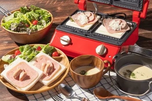 赤いホットサンドメーカーで調理中のホットサンドと、サラダ、ハムとソーセージのオープンサンド、スープが並んだ美味しそうな朝食またはブランチの食卓の様子です。温かい料理と新鮮な野菜がバランス良く配置されています。