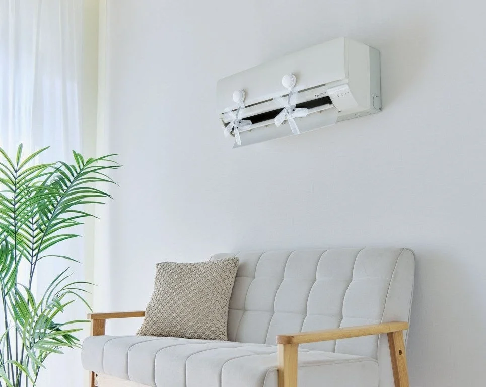 白い壁のリビングルームで、風よけカバーが取り付けられたエアコンの下に、シンプルなソファと観葉植物が配置されています