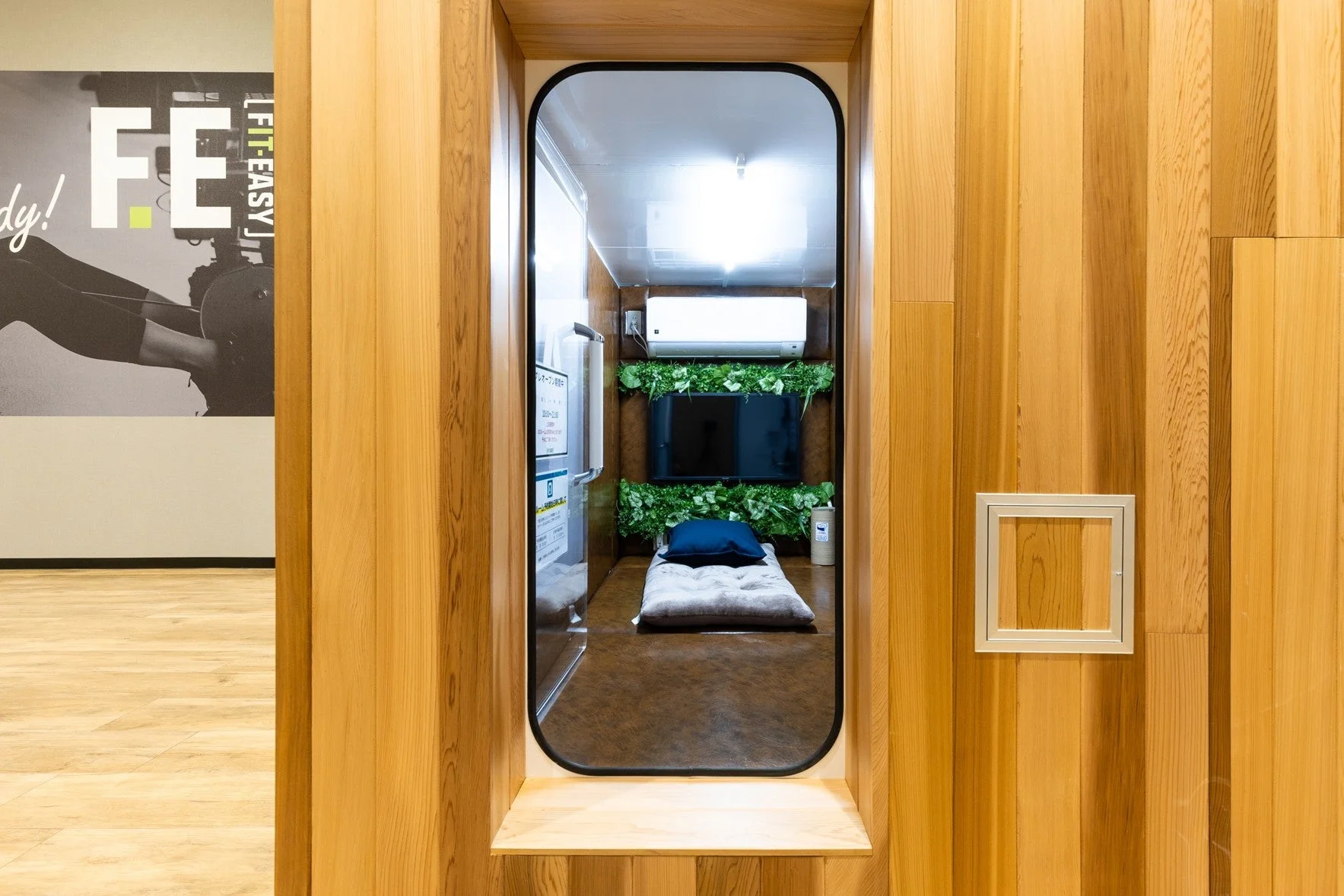 フィットネスジム内に設けられた木目調の個室リカバリールーム。テレビやエアコンが完備され、仮眠用の布団と青い枕が置かれています。壁にはジムのロゴが見えます。