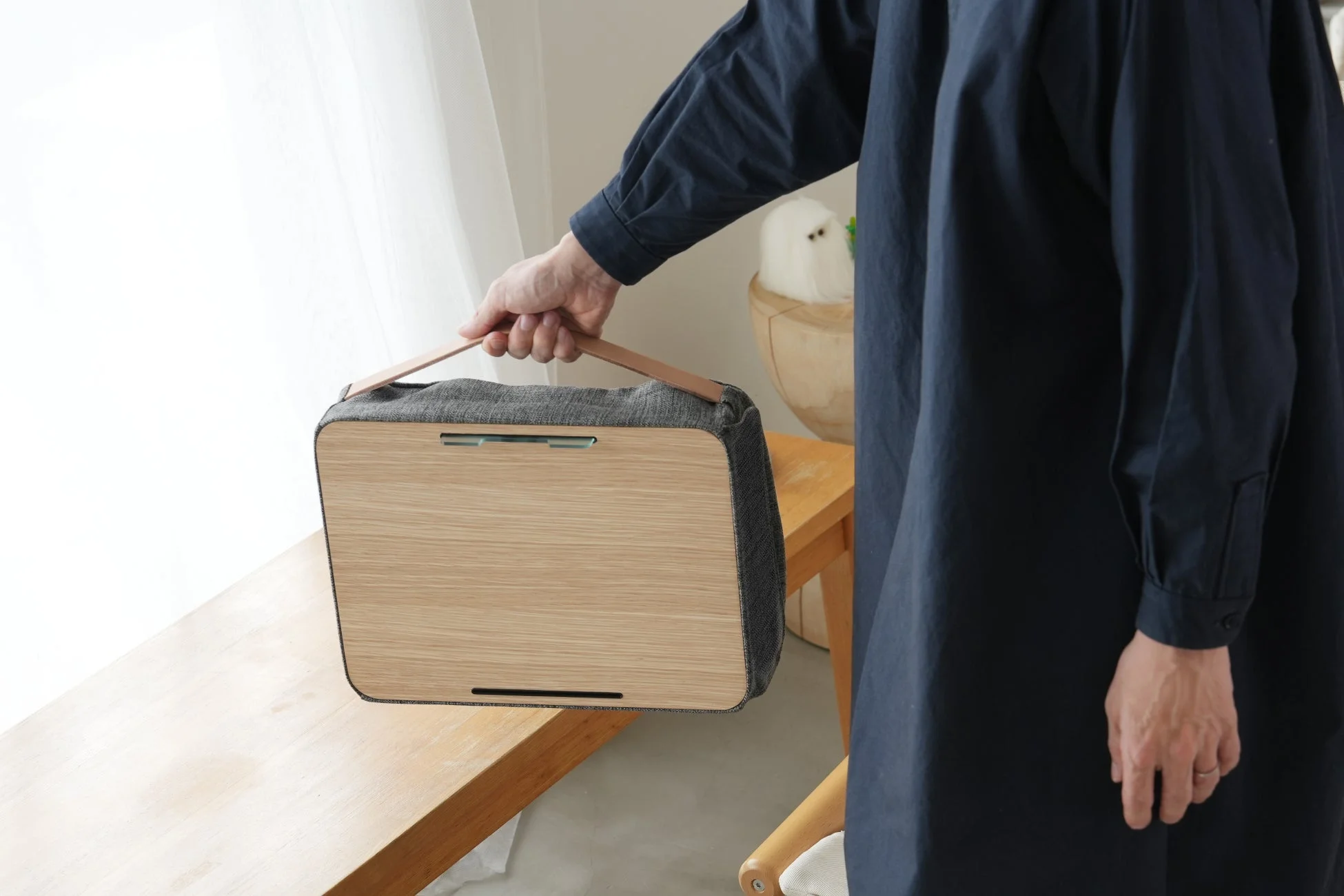 紺色の服を着た人物が、木目調の前面とグレーの側面、レザーの持ち手がついたポータブルデバイスを手に持っている様子。窓からの光が差し込む室内で、シンプルでモダンなデザインのガジェットが際立っています。