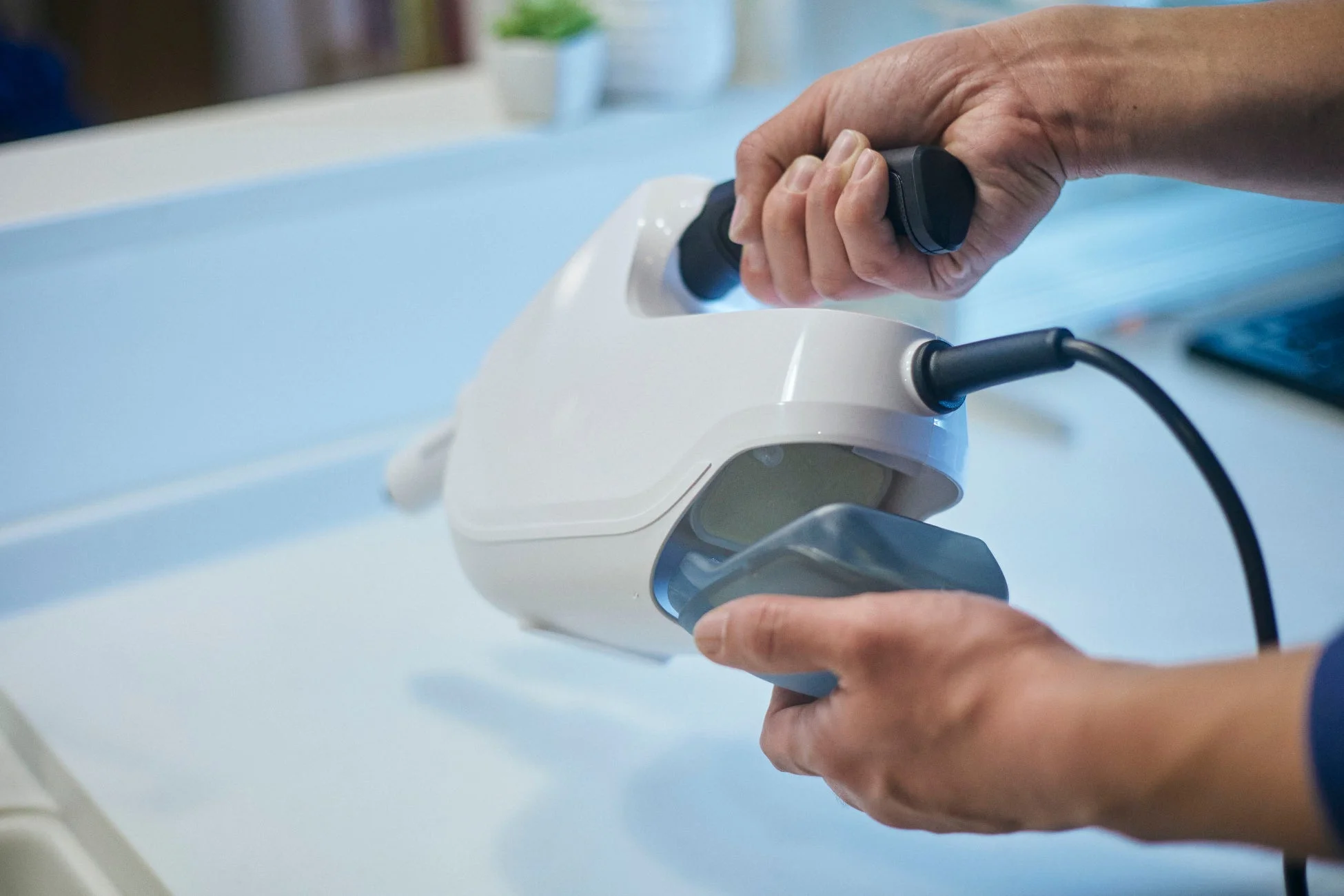 白いハンディデバイスを操作する人の手のクローズアップ。デバイスには黒いケーブルが接続されており、清潔な環境下での作業を示唆している。医療現場やラボでの使用が考えられる。