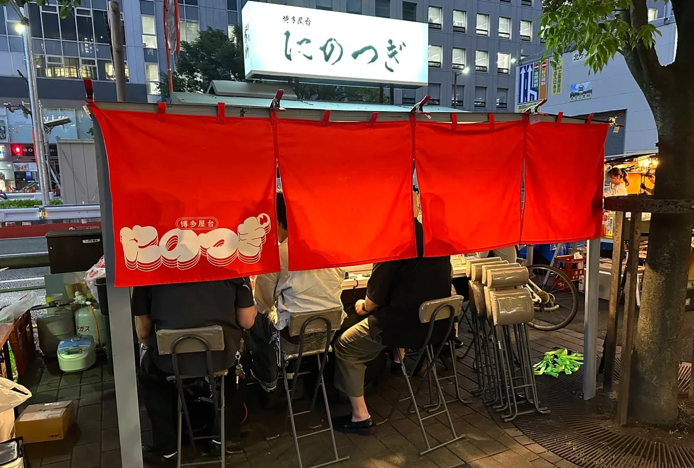 赤い暖簾がかかった博多屋台が夜の街角に佇む様子を捉えた画像です。数人の客がカウンター席で食事をしており、活気ある日本のストリートフード文化が感じられます。看板には「博多屋台 しにのつぎ」と書かれています。