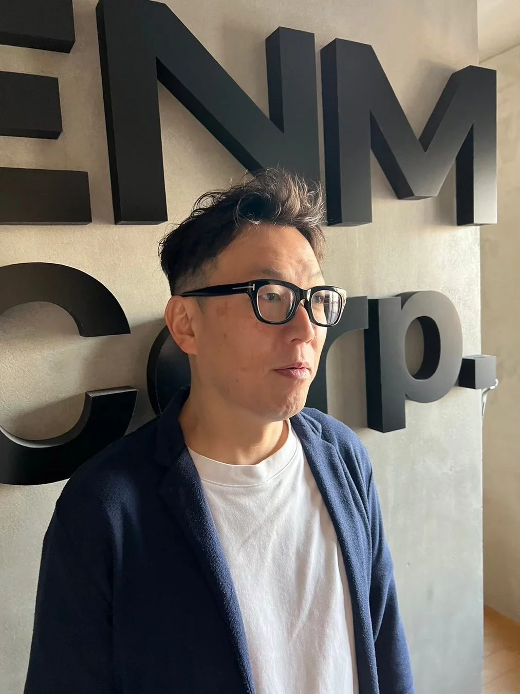 眼鏡をかけた男性がENM corp.と書かれた壁の前で横を向いている