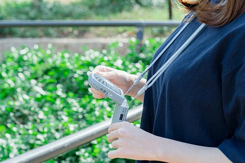 屋外で首から下げたポータブル扇風機を手に持つ人物の様子。暑い日に涼をとるためのガジェットを使用している場面を捉えています。