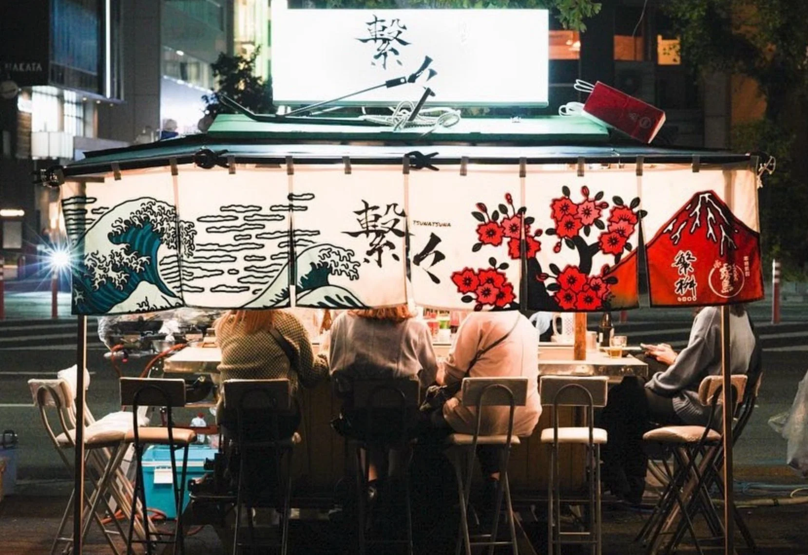 夜の街角に佇む和風の屋台で、人々がカウンターに座りくつろいでいる。暖簾には波、桜、赤富士といった日本の伝統的な模様が描かれ、賑やかな雰囲気が伝わる一枚。