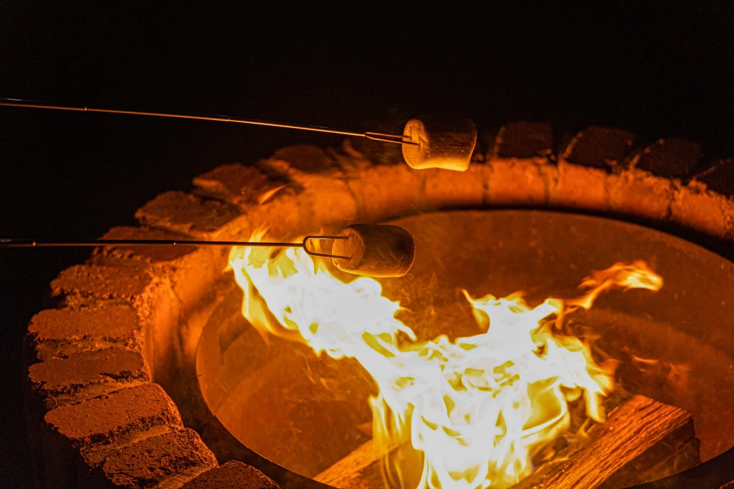 夜のファイヤーピットで、炎を囲んでマシュマロを焼いている様子を捉えた画像です。温かい光が心地よい、リラックスしたアウトドアの雰囲気が伝わってきます。