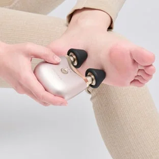 ベージュの衣服を着た人物が、小型の美容マッサージ器を素足の裏に当てて使用している様子