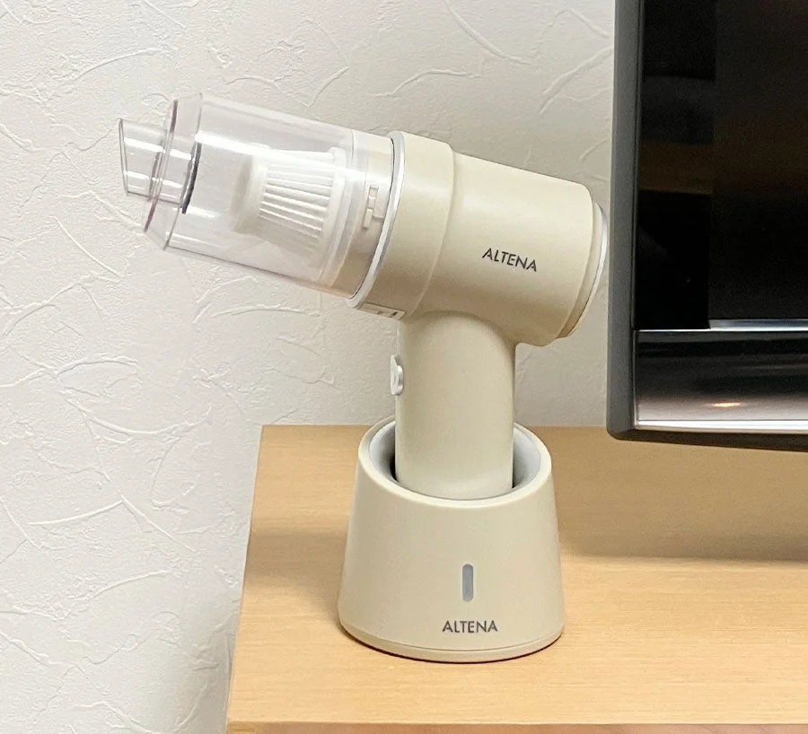 ベージュ色のハンディクリーナーが充電スタンドに置かれ、木製の台の上に設置されている様子。壁を背景に、スマートなデザインが特徴的な家電製品です。