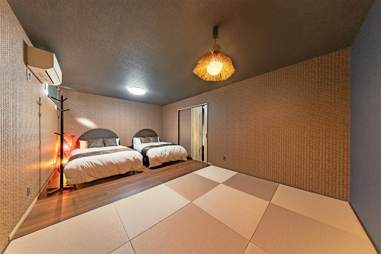 2つのベッドと畳スペースが設けられた、和モダンな雰囲気の寝室です。天井の独特な照明や壁の質感が特徴的で、落ち着いた滞在空間を提供しています。