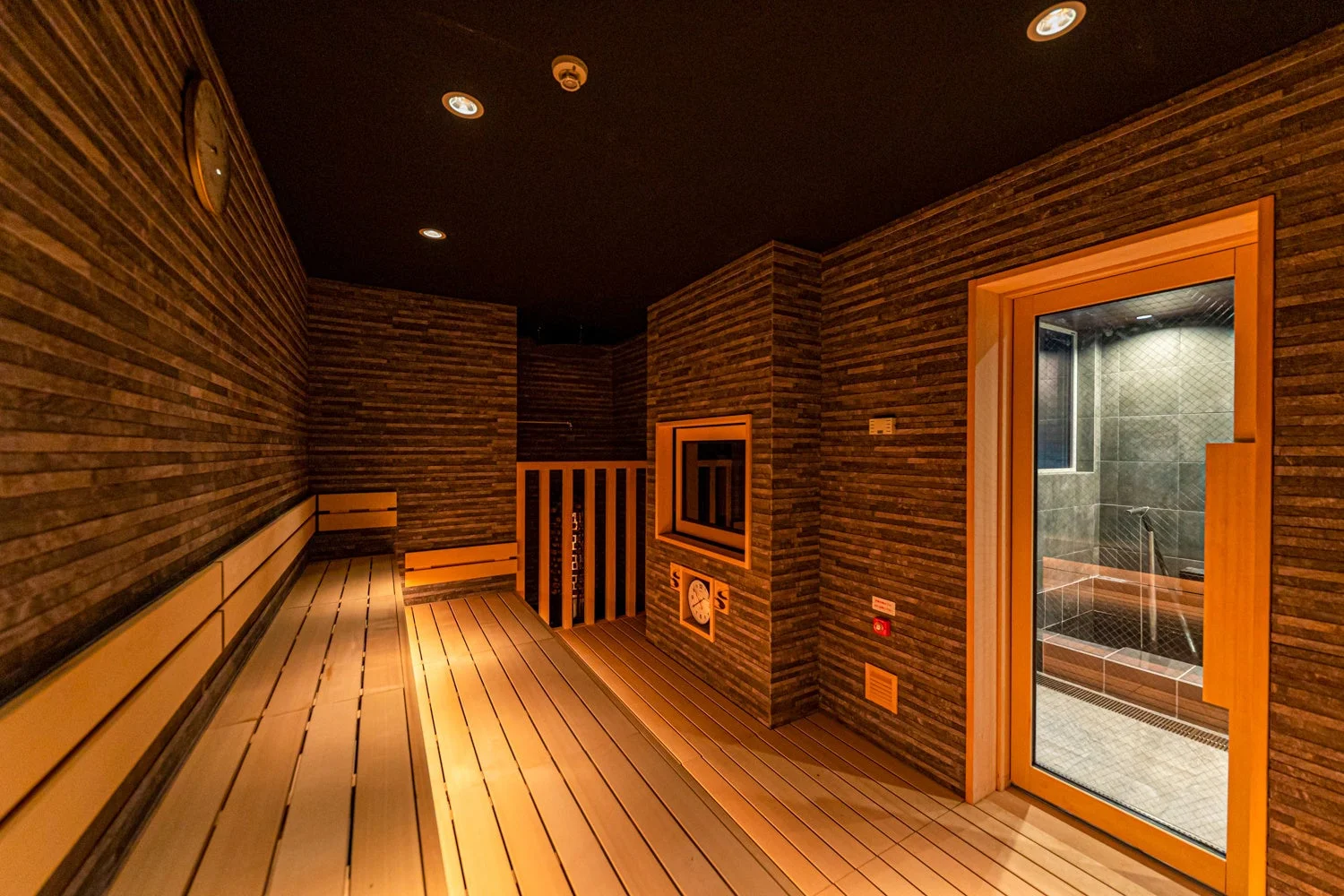 木製のベンチと壁が特徴的なモダンなサウナ室の全景。暖色系の照明が心地よい空間を演出し、奥には石造りの壁とガラス扉のシャワー・水風呂エリアが見える。清潔感があり、リラックスできる温浴施設の一部である。