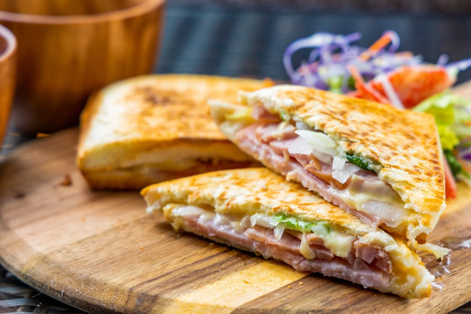 焼かれたホットサンドイッチが木製のカッティングボードに盛られています。ハム、とろけるチーズ、玉ねぎが挟まれ、手前のものは斜めにカットされています。奥にはもう一つ、右奥には付け合わせのサラダが見えます。